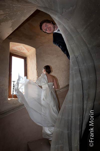 Les mariés dans un escalier du château, la mariée regarde par une fenêtre sa robe vole au vent