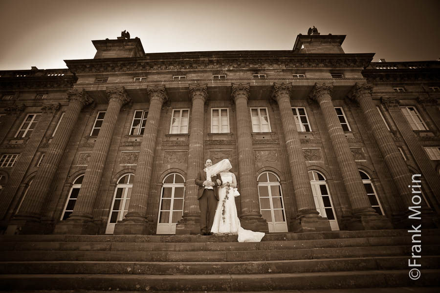 Photo au grand-angle en extérieur. Les Mariés côte-à-côte en haut d'un escalier. Derrière eux, les colones du château de Rohan à Saverne. Avec le vent, le voile cache le visage de la Mariée.