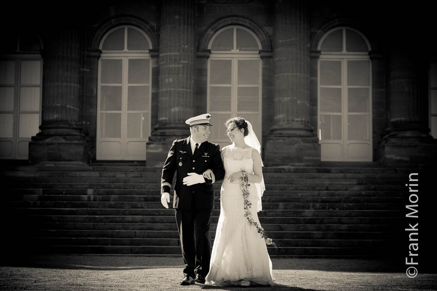 Photo au télé-objectif en extérieur. Les Mariés côte-à-côte se regardent en sourriant. Derrière eux, les colones du château de Rohan à Saverne.